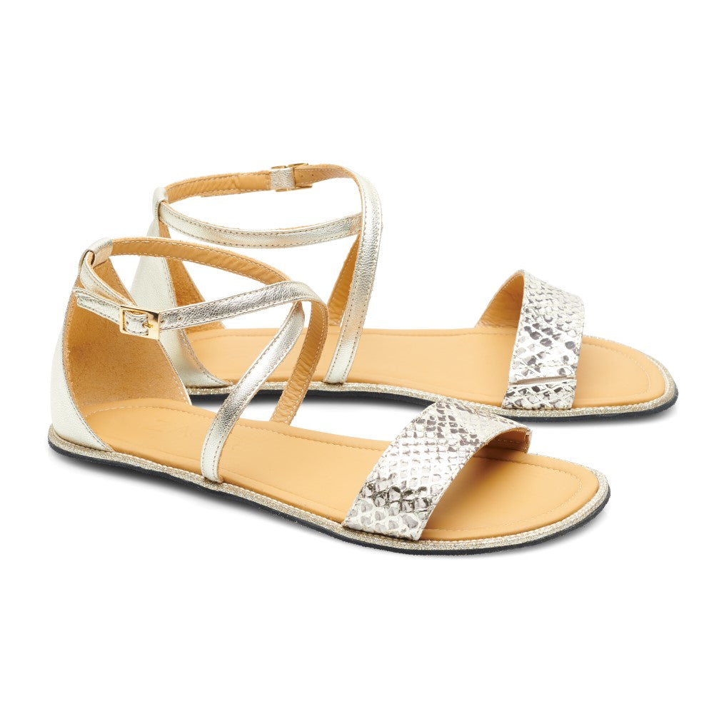 Wir präsentieren die QEEP Gold von ZAQQ: Diese flachen Sandalen für Damen sind vom Stil der Barfuß-Sandalen inspiriert und zeigen metallisch-silberne Riemen mit einem stilvollen Schlangenhautmuster. Sie verfügen über Knöchelriemen mit Schnallenverschluss und hellbraune Fußbetten, alles ergänzt durch elegante schwarze Sohlen auf schlichtem weißem Hintergrund.