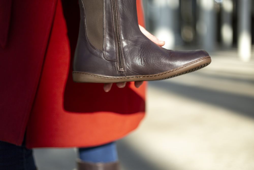 Eine Nahaufnahme zeigt eine Person in einem langen roten Mantel, die den ZAQQ QATCH Nappa Brown-Stiefel mit Reißverschluss hält. Der unscharfe Hintergrund lässt auf eine Außenumgebung schließen, in der Sonnenlicht Schatten wirft.