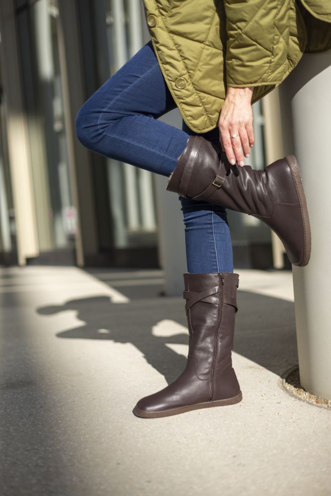 Eine Person in einem grünen Mantel und Blue Jeans lehnt an einer Säule und richtet einen ATTRAQT Winter Brown-Stiefel. Die Szene spielt im Freien, wobei das Sonnenlicht Schatten auf den Boden wirft, was den Komfort erhöht und dank des Designs von ZAQQ trotz der Kälte in der Luft ein Barfußgefühl vermittelt.