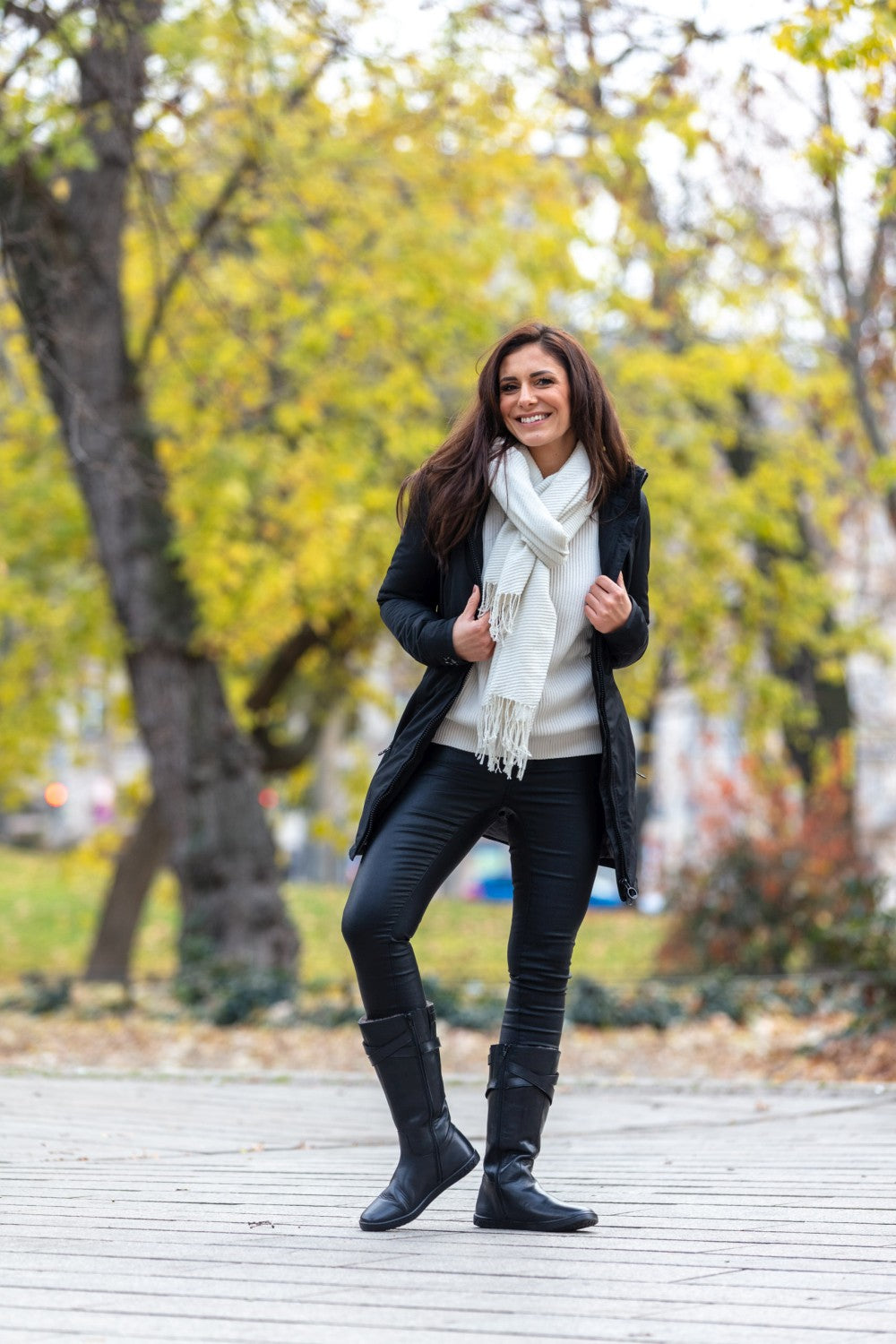 Eine Frau lächelt im Freien. Sie trägt einen schwarzen Mantel, einen weißen Schal und stylische ATTRAQT Winter Black-Stiefel von ZAQQ. Sie steht auf einem Holzweg, umgeben von Herbstbäumen und gefallenen Blättern, und strahlt eine fröhliche Herbstatmosphäre aus.