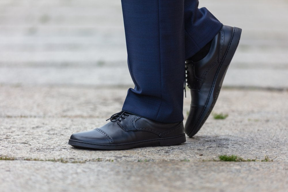 Eine Person in dunkelblauer Hose und den edlen BRIQ Captoe Black Schuhen von ZAQQ steht auf einem Steinpflaster. Die eleganten Schuhe im Brogue-Muster und mit hübscher Schnürung verleihen dem Look Raffinesse. Im Hintergrund runden leicht verschwommene Schritte die Szene ab.