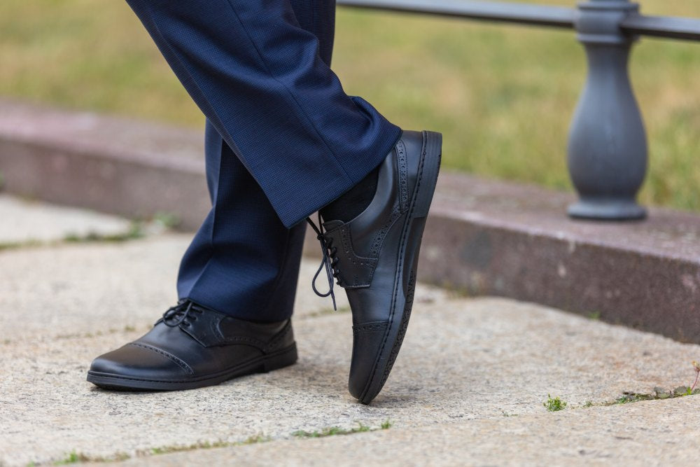 Eine Person in blauer Hose und dem stylischen BRIQ Captoe Black von ZAQQ steht auf einem Steinweg und lehnt im Hintergrund an einem unscharfen Zaun.