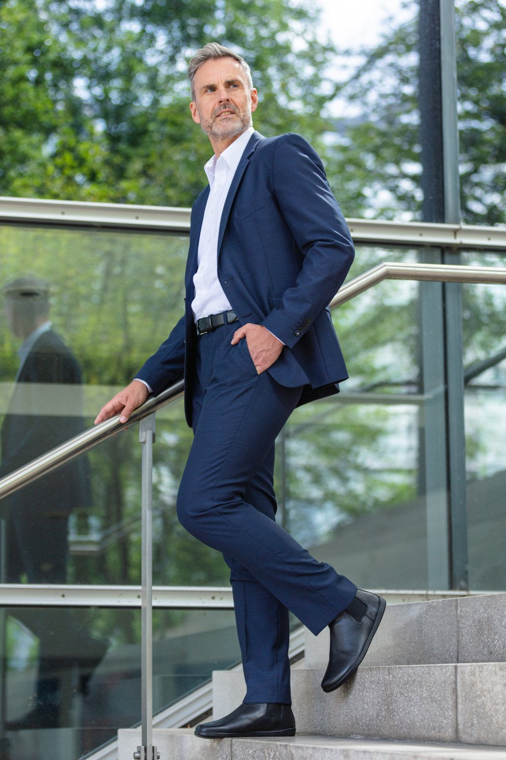 Ein Mann in marineblauem Anzug und weißem Hemd steht selbstbewusst auf einer Außentreppe und trägt die eleganten schwarzen EQUITY Brogue-Schuhe von ZAQQ. Eine Hand ruht in seiner Tasche vor einem Hintergrund aus Glasgeländern und üppigem grünem Laub, was eine professionelle und doch natürliche Atmosphäre schafft.