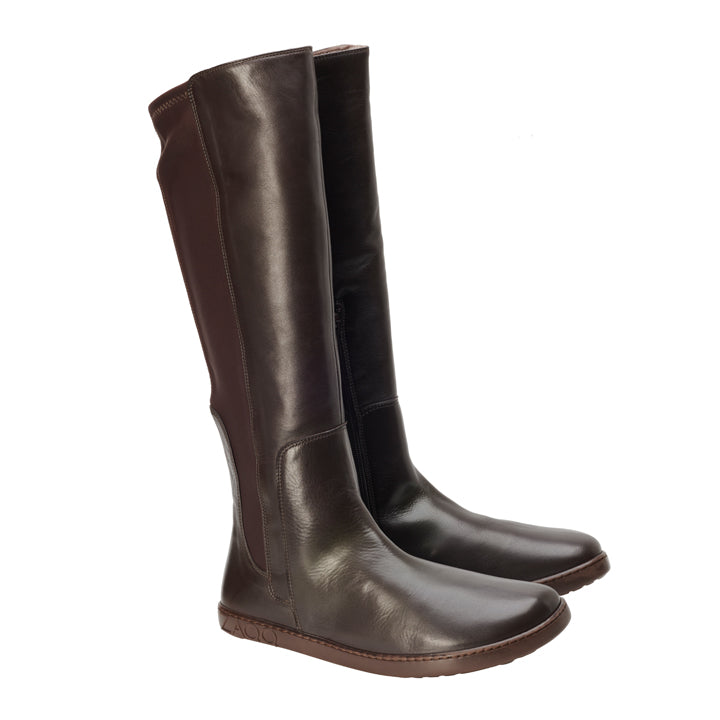 Die QATCH Winter Brown-Stiefel von ZAQQ sind elegant und zeichnen sich durch eine hohe Außenseite aus dunkelbraunem Nappaleder mit seitlichen Reißverschlüssen und flachen Sohlen aus, gefüttert mit kuscheligem Lammfell, alles aufrecht vor einem weißen Hintergrund.