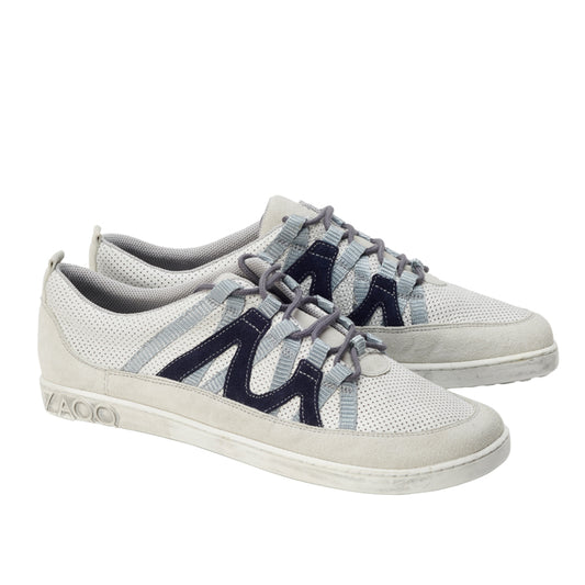 Ein Paar DRAQE White Grey Sneaker von ZAQQ aus Premium Nappaleder, mit perforierter Textur und dunkelblauen Zickzack-Streifen. Hellgraue Schnürsenkel betonen das Design und die flache Sohle weist ein geprägtes Branding an der Ferse auf. Die Schuhe sind von der Seite abgebildet und stehen für raffinierte Eleganz.