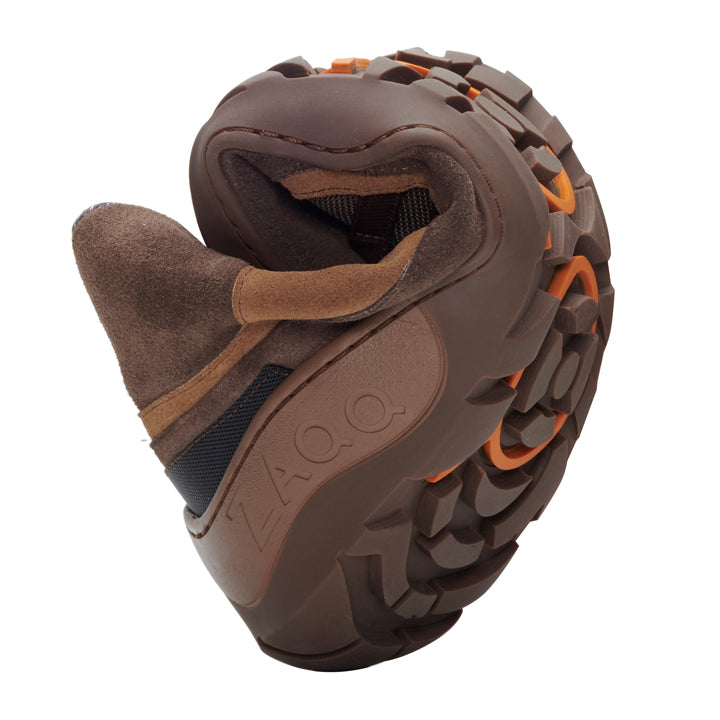 Der ZAQQ SQOUT Brown Waterproof ist ein vielseitiger Wanderschuh aus Veloursleder mit einer strapazierfähigen, strukturierten ZAQQ TERRA-Sohle und orangefarbenen Details. Wenn man den Schuh in der Mitte biegt, um seine Flexibilität hervorzuheben, ist an der Seite deutlich der Markenname ZAQQ zu sehen.