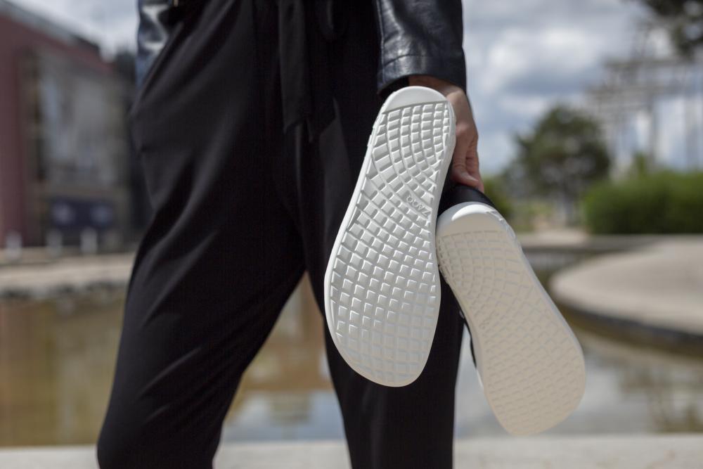 Eine Person in der schwarzen ALIQANTE-Kleidung von ZAQQ hält ein Paar Barfußschuhe mit strukturierten weißen Sohlen und steht im Freien an einer reflektierenden Wasseroberfläche mit einer verschwommenen Stadtlandschaft im Hintergrund.