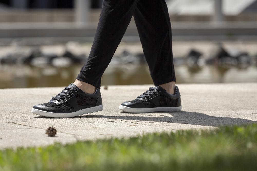 In einem Paar eleganter und atmungsaktiver schwarzer ZAQQ ALIQANTE-Sneakers schlendert eine Person einen Betonweg entlang. Im Vordergrund verleiht verschwommenes Gras der Szene Struktur, während ein von Felsen gesäumter Teich den Hintergrund ziert. Ihre schwarzen Hosen harmonieren nahtlos mit dem stylischen Schuhwerk.