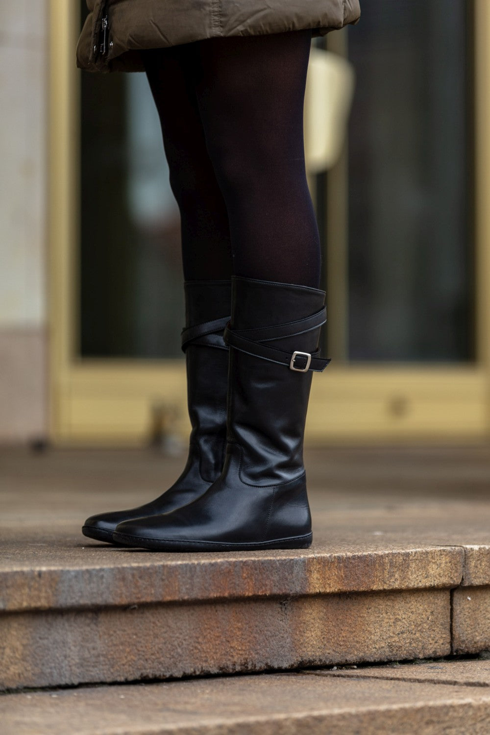 Nahaufnahme der Beine einer Person, die elegante schwarze Leggings und kniehohe ZAQQ ATTRAQT Nappaschwarze Stiefel mit Schnallen trägt und auf Steinstufen vor einer Glastür steht.