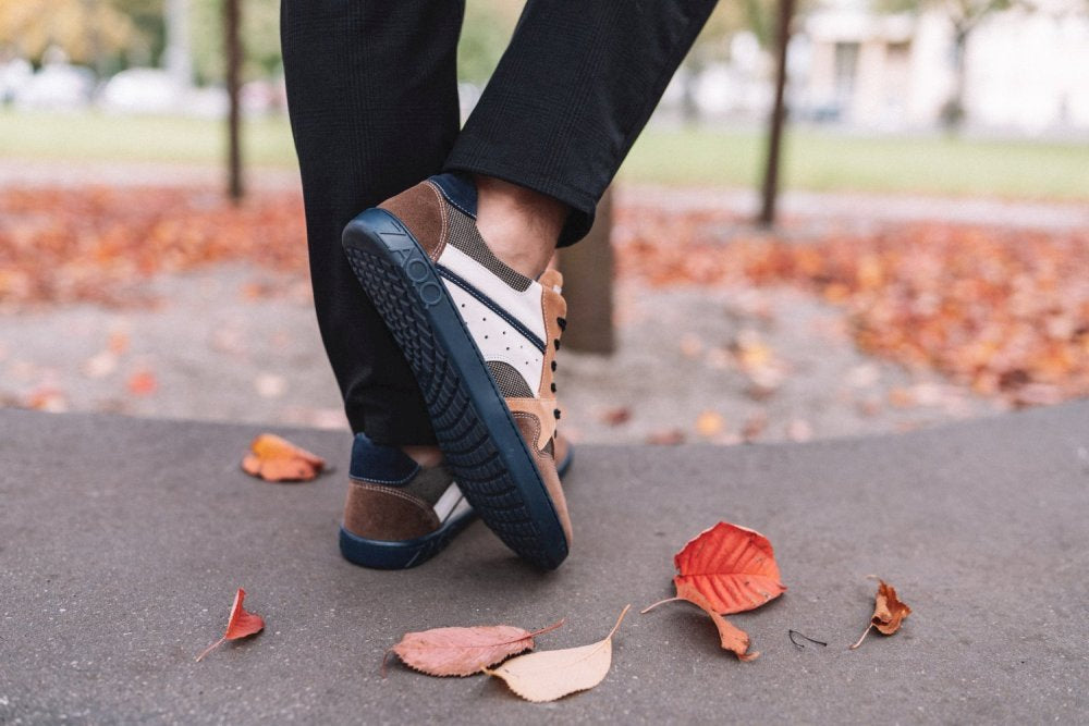 Eine Person in schwarzer Hose und schicken BREAQ Sierra Sneakers mit flexibler ZAQQ-Sohle spaziert einen mit Herbstlaub übersäten Weg entlang. Der Hintergrund ist unscharf und hebt Bäume und eine Grasfläche hervor.