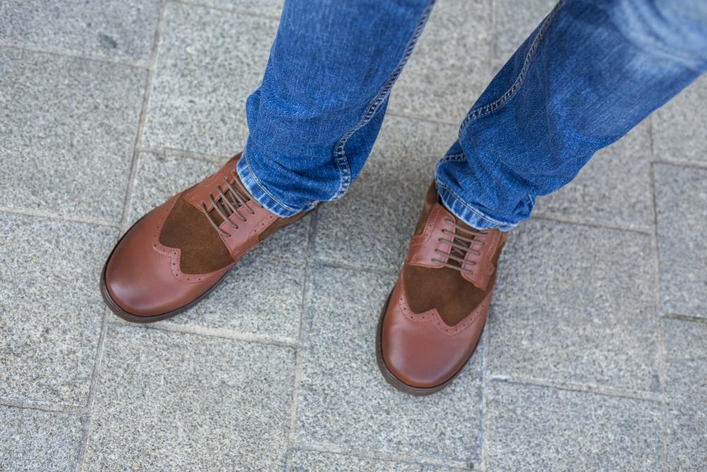 Eine Person in den Schuhen BRIQ Brogue Antique Cognac von ZAQQ steht auf einem grau gefliesten Bürgersteig. Sie trägt außerdem Blue Jeans.