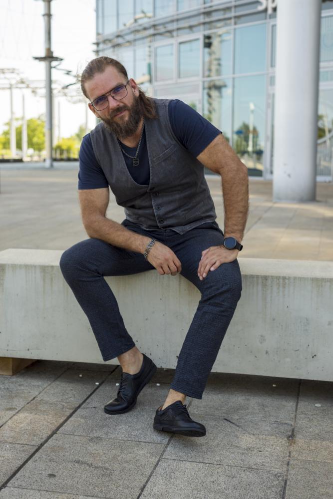 Ein bärtiger Mann mit langen Haaren sitzt draußen auf einer Betonbank. Er trägt eine Brille, ein blaues Hemd, eine graue Weste und eine blaue Hose. Seine Füße stecken bequem in den Schuhen BRIQ Brogue Tresse Black von ZAQQ. Im Hintergrund ergänzt ein modernes Gebäude die umliegenden Bäume.