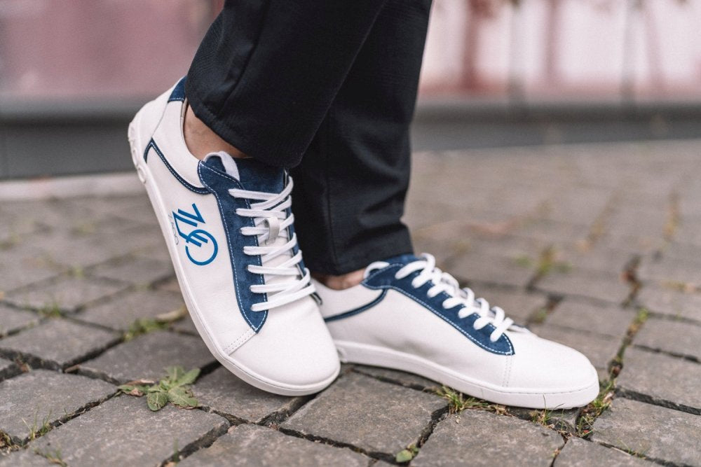 Eine Person, die ZAQQs CEDRIQ Blue Sneaker mit Fahrradlogo trägt, steht auf einem Kopfsteinpflaster. Diese Low-Top-Sneaker mit weißen Schnürsenkeln passen perfekt zu ihrer dunklen Hose.