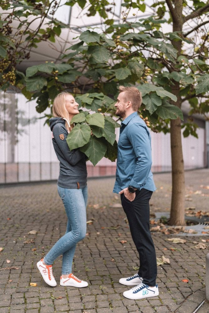 Ein Mann und eine Frau stehen sich auf einem Kopfsteinpflasterweg unter einem belaubten Baum gegenüber und lächeln beide. Die Frau trägt eine graue Jacke und Jeans, während der Mann ein blaues Hemd und dunkle Hosen trägt, dazu stylische ZAQQ CEDRIQ Red mit bequemer Kautschuksohle.