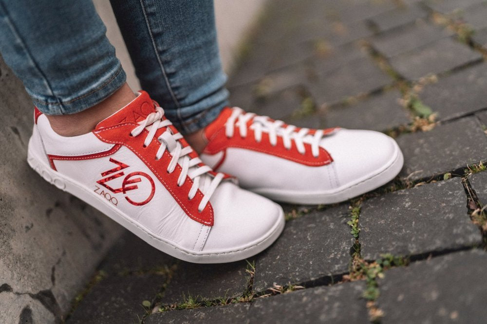 Eine Person, die die CEDRIQ Red Sneaker von ZAQQ trägt, steht auf einer Kopfsteinpflasterfläche. Diese stylischen Barfuß-Sneaker haben eine weiße Farbe mit roten Akzenten und Logo, verfügen über Schnürsenkel und ein seitliches Design sowie eine bequeme Kautschuksohle. An den Unterschenkeln sind blaue Jeans zu sehen.