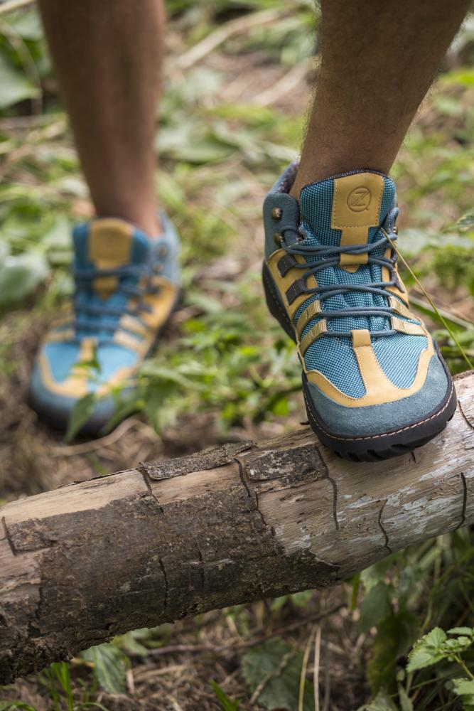 Eine Person, die die wasserdichten Wanderschuhe ZAQQ ESQAPE Blue Orange trägt, tritt in einer Waldlandschaft auf einen umgestürzten Baum. Der Hintergrund ist mit üppigem grünem Laub und Gras gefüllt und schafft eine bezaubernde Atmosphäre.