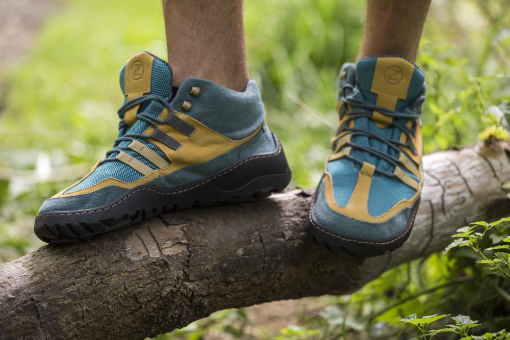 Eine Person in blauen und orangefarbenen ZAQQ ESQAPE-Schuhen steht auf einem Baumstamm in einer Waldlandschaft. Diese Wanderschuhe haben dicke, wasserdichte Sohlen und ein Schnürdesign, was sie ideal für Outdoor-Aktivitäten macht. Das umgebende grüne Laub verstärkt das Gefühl von Abenteuer.