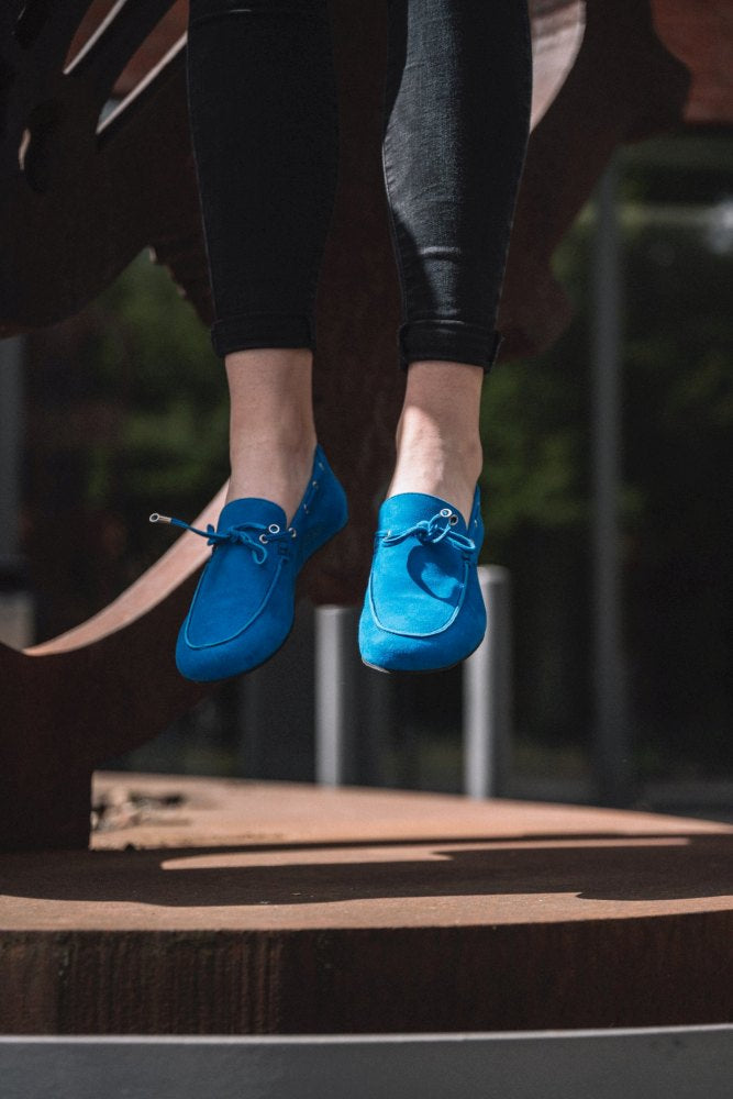 Ein Paar Füße in leuchtendem Blau von FLIQ Blue von ZAQQ schwebt in der Luft. Die Person trägt dunkle Leggings und befindet sich im Freien vor einem verschwommenen Hintergrund aus Grünpflanzen und architektonischen Elementen.