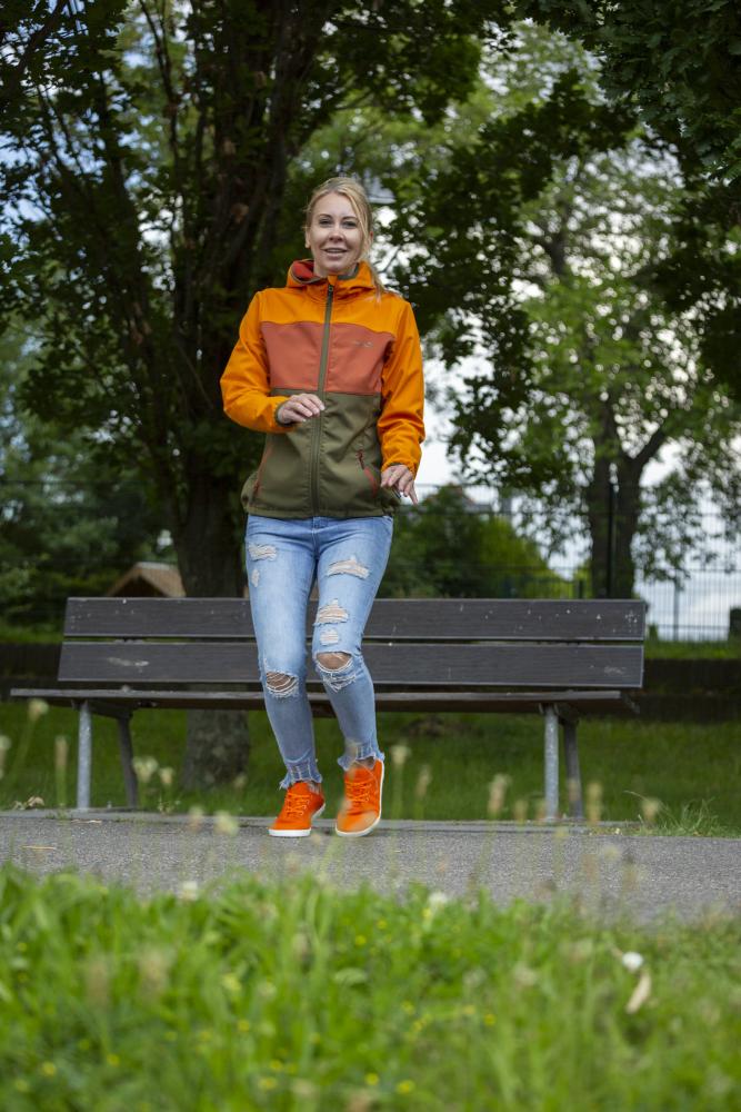 Eine Frau in orange-grüner Jacke und zerrissenen Jeans läuft auf einem Weg im Park und genießt das Barfußgefühl, das ihre leuchtend orangefarbenen LIQE-Schuhe von ZAQQ vermitteln. Lächelnd geht sie unter einem bewölkten Himmel an einer Holzbank und grünen Bäumen vorbei.