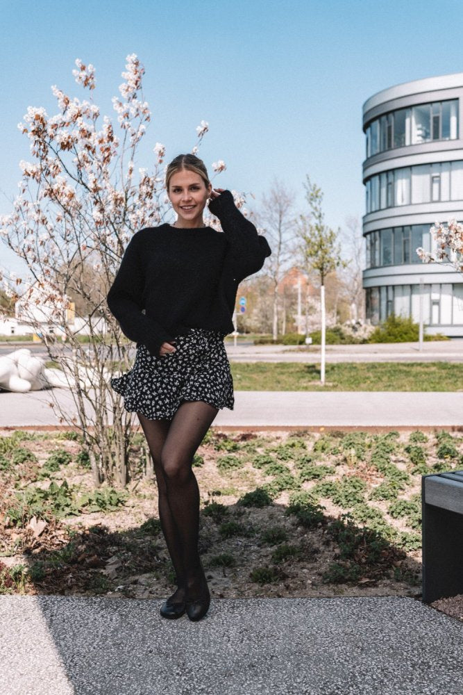 Eine Person steht lächelnd im Freien und trägt einen Pullover und einen geblümten Rock aus der ZAQQ-Kollektion. Das Outfit im Farbton LOOQ Dark Brown trägt zu ihrem mühelosen Stil bei, der an eine Barfuß-Ballerina erinnert. Sie werden von blühenden Bäumen und einem modernen Gebäude im Hintergrund unter dem klaren blauen Himmel eingerahmt.