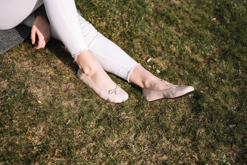 Eine Person in weißer Hose und LOOQ Nude Ballerinaschuhen von ZAQQ sitzt mit ausgestreckten Beinen auf grünem Gras und strahlt Eleganz aus. Die Sonne wirft weiche Schatten auf das Gras und schafft eine heitere und gemütliche Szene.