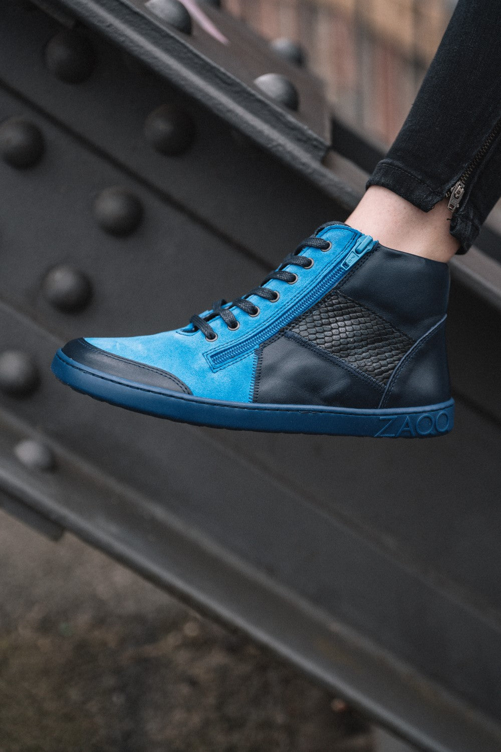 Eine Nahaufnahme eines stylischen azurblauen und schwarzen High-Top-Sneakers aus der LUQQ Azur-Kollektion von ZAQQ, getragen von einer Person, die auf einem Metallträger steht. Der Sneaker weist ein markantes Strukturmuster auf, wobei das ZAQQ-Logo prominent auf der Zwischensohle abgebildet ist. Die Person trägt dunkle Hosen.