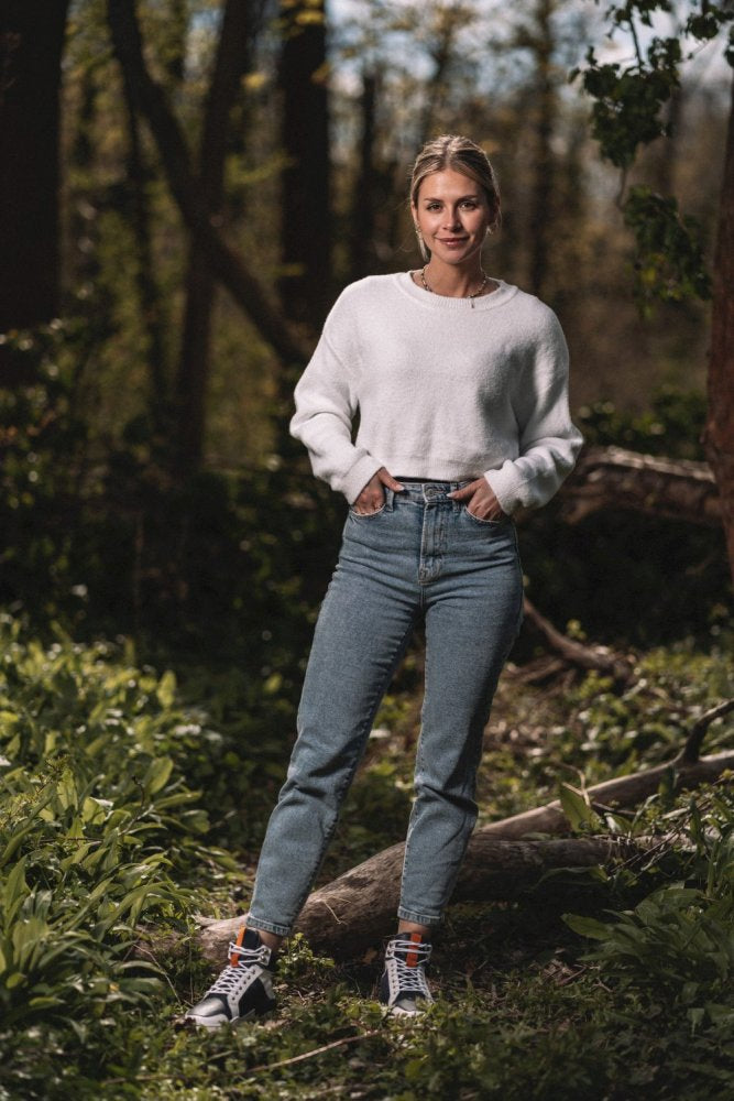 Eine Person steht in einem Wald und trägt einen weißen Pullover, blaue Jeans und High-Top-Sneakers von ZAQQ mit dem MONTBLANQ Blue White Waterproof-Design. Im Hintergrund sind Bäume und üppiges Grün zu sehen, durch das Sonnenlicht fällt. Die Person hat die Hände in den Taschen und lächelt inmitten der Natur.