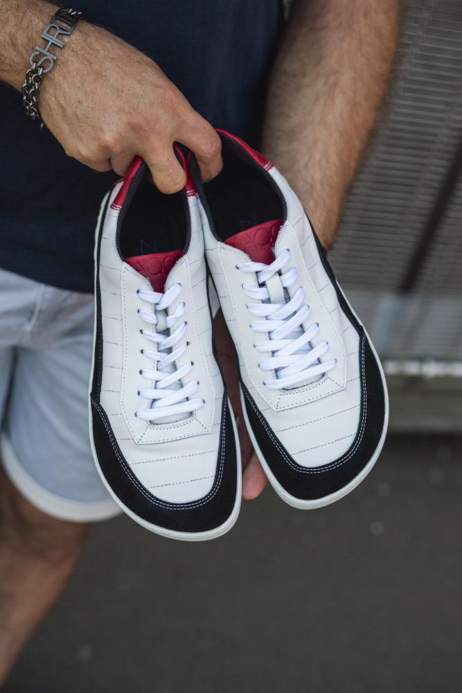 Eine Person hält ein Paar ZAQQ QAMPION Black White Sneaker, mit rotem Innenfutter und schwarzen Details. Die flexible Sohle erhöht den Tragekomfort. Sie trägt ein dunkles Shirt mit hellgrauen Shorts, wobei der Fokus auf den stylischen Sneakern liegt.