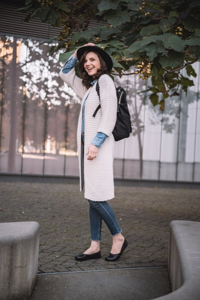 Eine lächelnde Frau steht draußen in der Nähe eines Gebäudes. Sie trägt eine lange weiße Strickjacke und blaue Jeans. Ihr Outfit komplettiert sie mit dunkelbraunen Ballerinas von ZAQQ QARMA. Ihr schwarzer Hut passt gut zu dem schwarzen Rucksack, den sie trägt, während sie ihn vorsichtig zurechtrückt. Große grüne Blätter hängen über ihr und verleihen ihrer ruhigen Pose einen Hauch von natürlichem Charme.