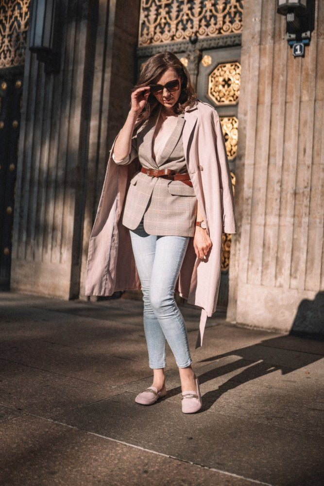 Eine Frau mit Sonnenbrille steht auf einer sonnenbeschienenen Straße. Sie trägt einen langen Mantel über den Schultern, einen Blazer mit Gürtel, helle Jeans und QERP Candy-Loafer von ZAQQ. Sie rückt ihre Sonnenbrille zurecht, im Hintergrund sind verzierte Türen und Steinmauern zu sehen.