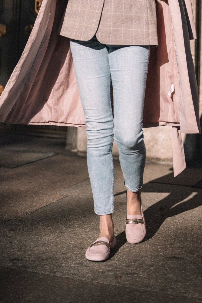 Eine Person in einem hellrosa Mantel, kariertem Blazer, hellblauen Röhrenjeans und ZAQQs QERP Candy Veloursleder-Loafern mit metallischen Akzenten läuft auf einem sonnenbeschienenen Bürgersteig. Das Bild fängt die untere Körperhälfte in einem stilvollen, lässigen Outfit ein.