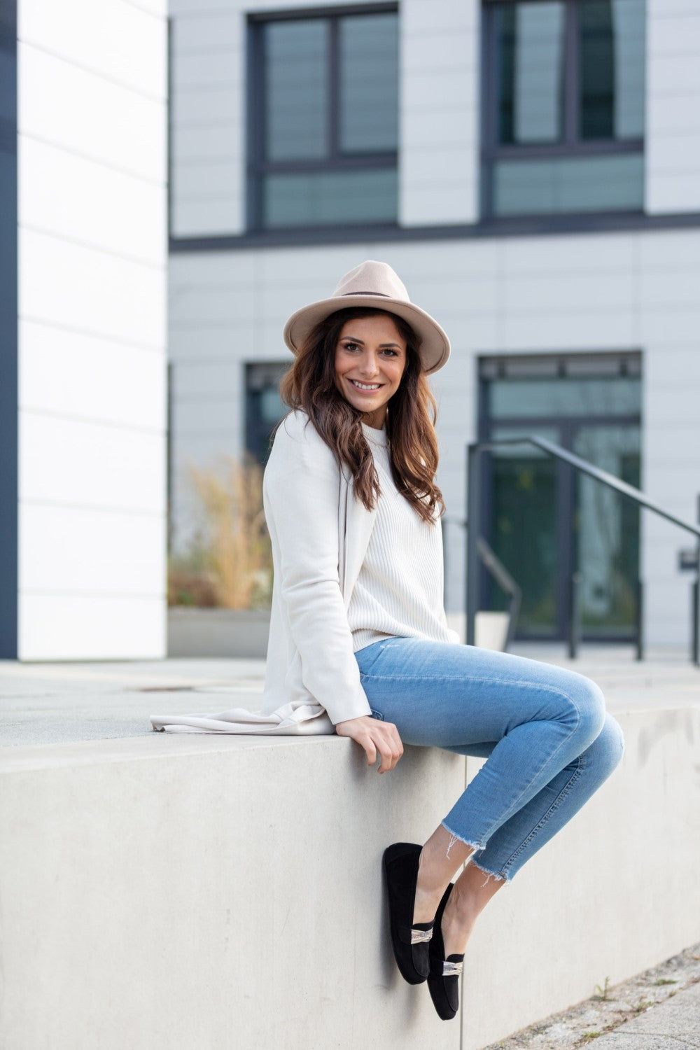 Eine Frau in weißer Bluse, heller Strickjacke und Jeans sitzt auf einem Felsvorsprung. Sie trägt einen schicken rosa Hut und ihre schwarzen ZAQQ QERP Velours-Schuhe. Sie lächelt in die Kamera vor dem Hintergrund eines modernen Gebäudes mit großen Fenstern.