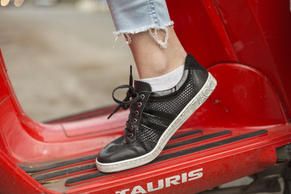 Eine Person in Barfuß-Sneakern mit weißer Sohle und weißen Söckchen steht auf der Fußstütze eines roten Rollers. Der Saum ihrer QIMBERLY Blue Jeans von ZAQQ ist ausgefranst, was dem Outfit einen lässigen Touch verleiht.