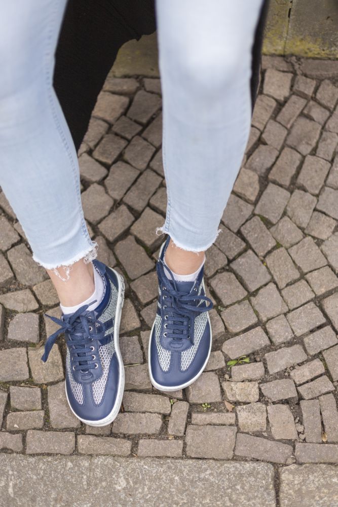 Eine Person in der Jeans QIMBERLY Blue von ZAQQ und blauen Barfuß-Sneakern aus hochwertigem Nappaleder steht auf einem Kopfsteinpflaster.