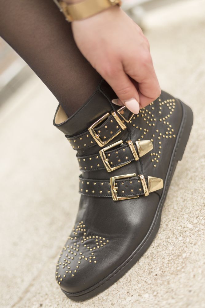 Eine Trägerin der QLANCE Black Gold Stiefeletten von ZAQQ rückt ihr stylisches Schuhwerk zurecht. Die eleganten goldenen Nieten und drei raffinierten goldenen Schnallen harmonieren perfekt mit dem filigranen Muster auf schwarzen Strumpfhosen und verkörpern eine schicke schwarz-goldene Ästhetik.