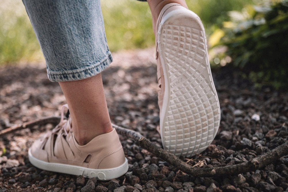 Eine Person, die QLEAR Light Pink-Sneakers von ZAQQ und Blue Jeans trägt, tritt auf einen kleinen Ast, während sie auf einem Kiesweg läuft. Die Sohle eines Sneakers ist sichtbar und weist ein detailliertes Profilmuster auf, das hervorragenden Halt bietet. Im Hintergrund ist Grün zu sehen.