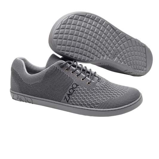 Ein Paar elegante vegane Sportschuhe QNIT Grey von ZAQQ mit atmungsaktivem Obermaterial aus Baumwollstrick und passenden grauen Sohlen. Ein Schuh steht aufrecht und hebt seine gemusterte Oberfläche und Schnürsenkel hervor, während der andere geneigt ist, um das Profilsohlendesign zu zeigen.