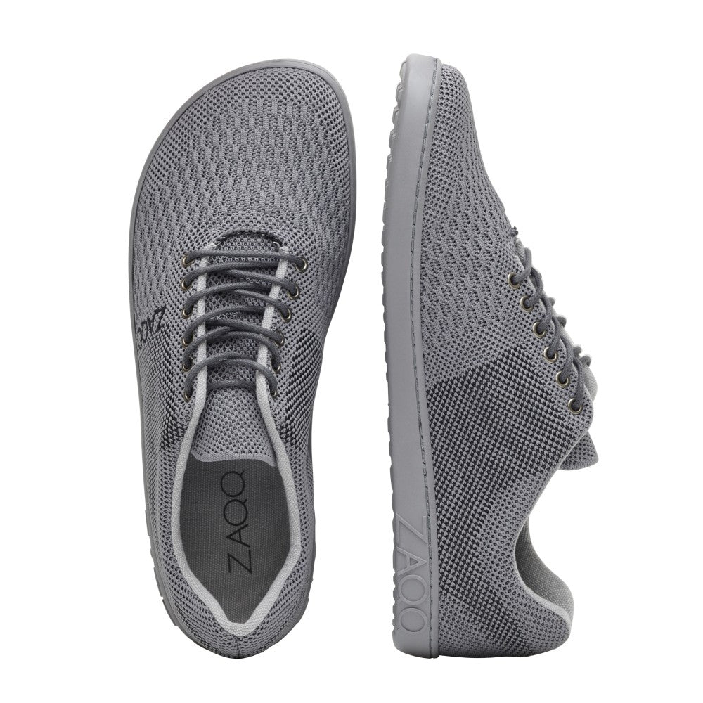 Ein Paar QNIT Grey-Sneaker von ZAQQ, von oben und von der Seite abgebildet, besteht aus einem atmungsaktiven Baumwollstrickgewebe. Diese veganen Schuhe verfügen über eine strukturierte Oberfläche mit flachen Gummisohlen und sorgen durch ihr Barfußschuhdesign für Komfort. Der Markenname ZAQQ ist sowohl an der Seite als auch auf der Innensohle deutlich sichtbar.