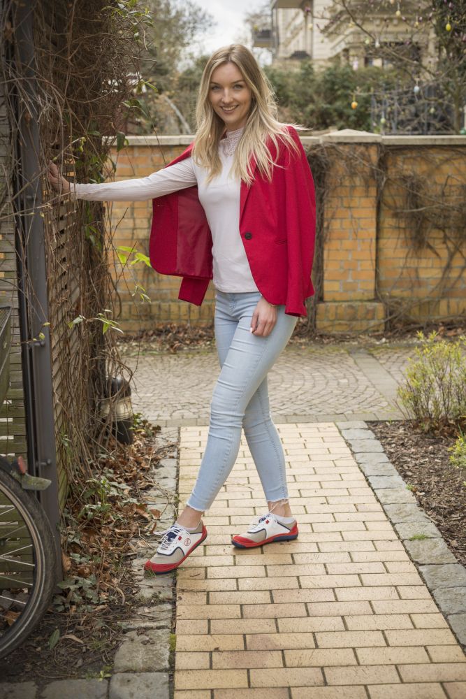 Eine Frau mit langen blonden Haaren, bekleidet mit einer roten Jacke, einem weißen Hemd und hellblauen Jeans, steht auf einem Ziegelweg. Sie lächelt und stützt eine Hand auf einen Holzzaun. Sie präsentiert die weißen QOLETTE-Sneaker aus der Kollektion der Marke ZAQQ. Im Hintergrund sind leuchtendes Grün und eine Ziegelwand zu sehen.