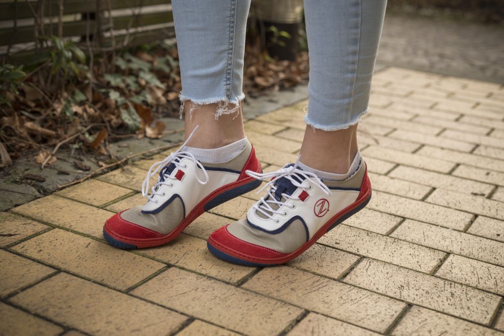 Eine Person, die die weißen ZAQQ QOLETTE-Sneakers trägt, schlendert einen gelben Ziegelweg entlang. Die Sneaker sind mit Schnürsenkeln versehen und das Ensemble wird durch hellblaue Jeans mit ausgefransten Säumen vervollständigt. Braune Blätter und Grünpflanzen verschönern die Hintergrundlandschaft.