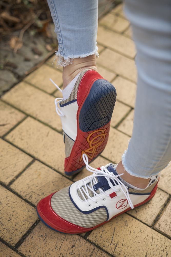 Eine Person trägt die leuchtenden weißen QOLETTE-Sneaker von ZAQQ mit roten, weißen und blauen Akzenten und steht auf einem Ziegelweg. Sie hat diese Sneaker mit hellblauen Jeans kombiniert, die an den Knöcheln leicht ausgefranst sind.