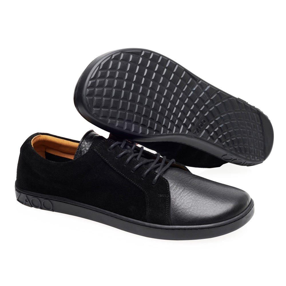 Präsentiert wird ein Paar ZAQQ QORE Low Black Schnürschuhe. Ein Schuh liegt auf der Seite und zeigt die Sohle mit Waffelmuster, während der andere aufrecht steht und die glatte Leder- und Wildlederstruktur zur Schau stellt.