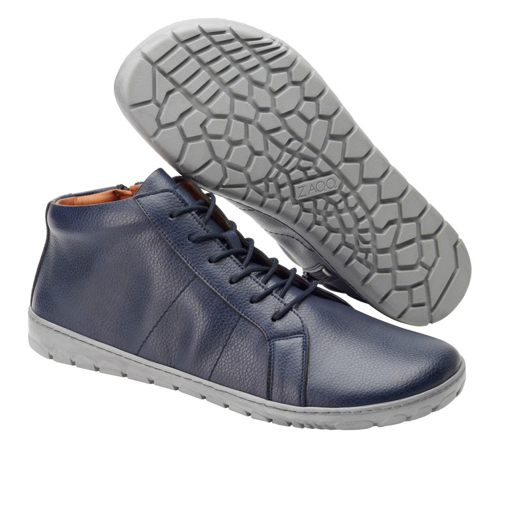 Wir präsentieren den QORE Mid Vegan Navy von ZAQQ: Ein stilvolles Paar marineblauer High-Top-Sneaker mit grauer Sohle. Diese Schuhe haben ein Obermaterial aus strukturiertem Leder und sind sowohl von der Seite als auch von unten zu sehen, wodurch das Profilmuster auf der flexiblen Sohle betont wird.