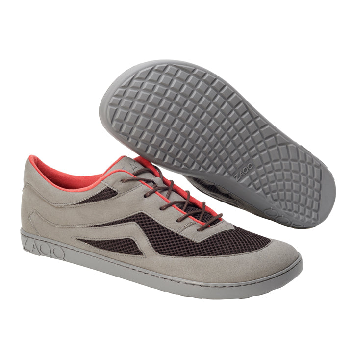 Die ZAQQ QYNLEE Grey Sneaker bestechen durch eine schicke Kombination aus Grau und Schwarz, ergänzt durch leuchtend rote Akzente. Ein Schuh steht aufrecht und zeigt das hochwertige Seiten- und Obermaterial aus Veloursleder, während der andere sein strukturiertes Sohlenmuster hervorhebt und sein vom Barfußlaufen inspiriertes Design betont.