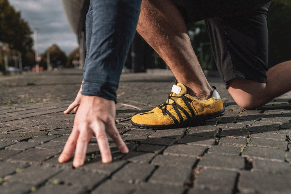 Eine Nahaufnahme zeigt eine Person in Startposition auf einem Ziegelsteinpflaster. Sie trägt die RIQ Yellow Laufschuhe von ZAQQ und schwarze Shorts. Ihre Hände sind auf dem Boden, bereit zum Sprinten, was ein flexibles und barfußähnliches Gefühl vermittelt. Der unscharfe Hintergrund gibt subtil Einblicke in Bäume und Himmel.