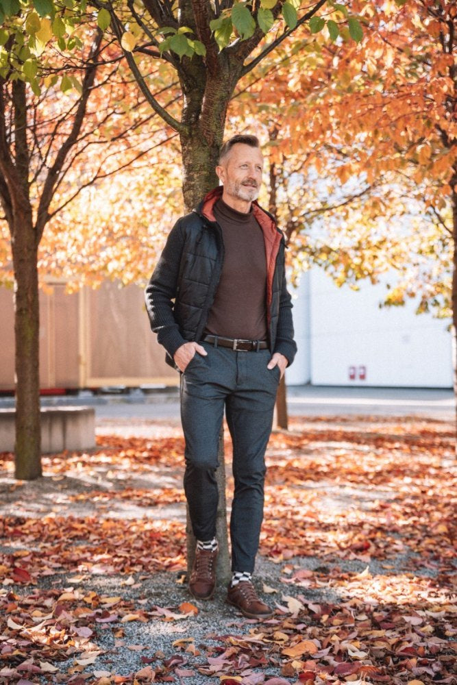 Ein Mann mit Bart steht neben einem mit Herbstlaub geschmückten Baum und präsentiert Tragekomfort in seinem Ensemble aus schwarzer Jacke und braunem Pullover. Er kombiniert es stilvoll mit grauen Hosen und RIQO Brown von ZAQQ für einen mühelos lässigen Look im Park, wo gefallene Blätter den Boden vor einer Kulisse aus Bäumen bedecken.