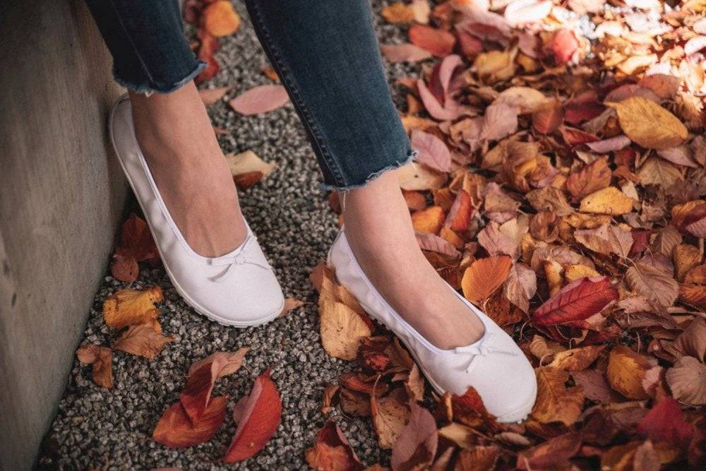 Eine Person steht auf einem Kiesweg, umgeben von gefallenen Herbstblättern in Rot-, Orange- und Brauntönen und trägt die weißen Ballerinas SUQAR mit Sneakersohle von ZAQQ und dazu dunkle Jeans.