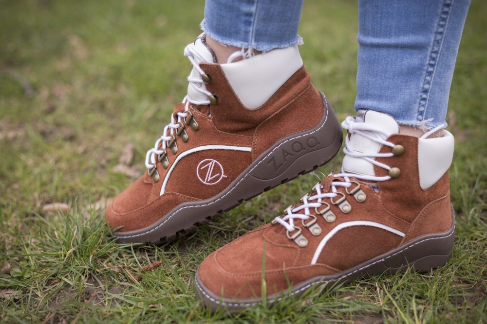 Eine Person, die die wasserdichten High-Top-Sneakers TREQ Cognac mit weißen Details und Schnürsenkeln trägt, steht auf Gras. Diese Outdoor-Schuhe von ZAQQ zeigen das Markenlogo an der Seite und die Person trägt Blue Jeans.