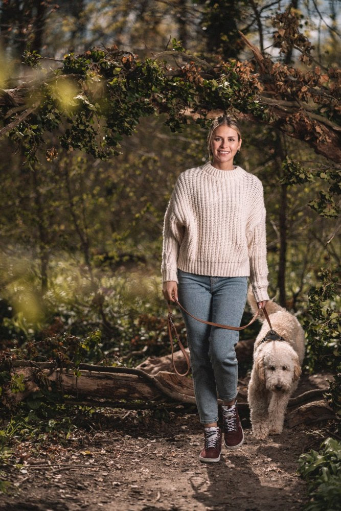 In den bequemen TREQ Red Waterproof-Schuhen von ZAQQ schlendert eine Person in cremefarbenem Pullover und Jeans mit ihrem flauschigen weißen Hund durch den Waldweg. Die Sonne scheint durch die Bäume und hebt das grüne Laub hervor.