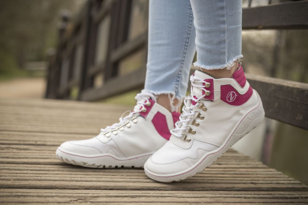 Eine Person, die die VAQATION Pink Waterproof Sneakers von ZAQQ trägt, steht auf einer Holzbrücke und trägt hellblaue zerrissene Jeans. Der Hintergrund ist leicht verschwommen und suggeriert eine Außenkulisse mit viel Grün.